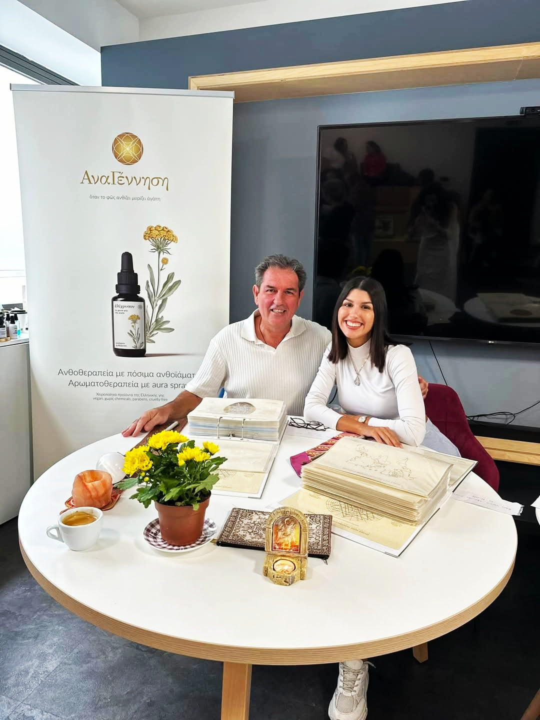 Art of Iasis Seminar Level 3 in Athens 2025 – Advanced holistic therapy workshop. Two people sitting at a table with books and a plant, with a branded backdrop in the background.